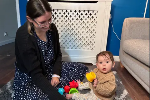 Chloe Chloe sits on a grey and cream rug with her daughter who is playing with colourful stacking cups and looking over at the camera, wearing a knitted beige jumper and leggings with a floral pattern. Chloe is wearing a black and white dress with a black cardigan, smiling down at her daughter.