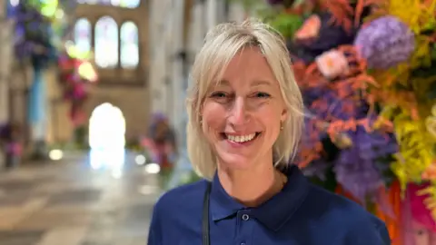 Caroline Daines who's wearing a navy blue polo top and has blonde hair standing in the main hall of the cathedral with a floral arrangement next to her