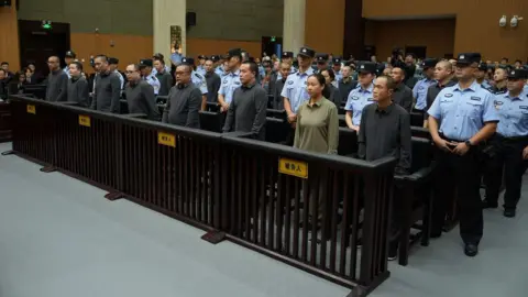 CCTV Seven men and a woman, dressed in grey and beige shirts stand in a row in a court, surrounded by security officers in blue shirts with black caps