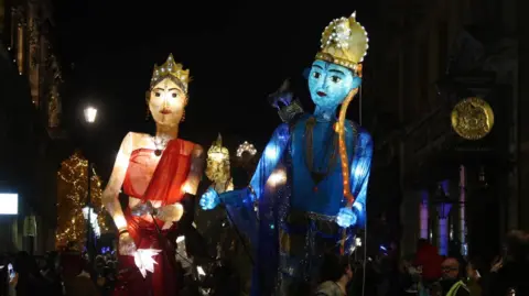 Diwali celebrations in Northampton on 26 October 2024. Featured are two giant puppets lit up with lights being carried down a dark street, brimmed with people. 