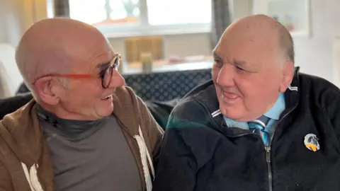 Photo of two men - support worker Ian Meikle and Tony, who has learning difficulties. They are  in a living room, sitting on a sofa, looking towards each other. Ian is bald and wears glasses with a orange/brown frame. Ian is also almost bald. He is wearing a blue shirt and striped tie under a zip-up cardigan.