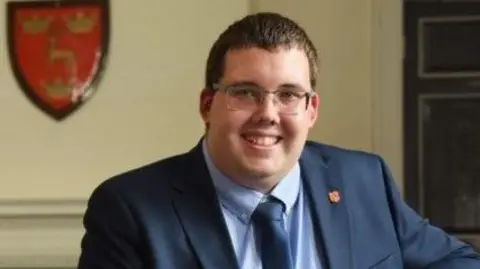 Dereham Town Council Stuart Green is in the middle. He has short brown hair and is wearing glasses, a blue shirt, blue tie and navy suit jacket. 