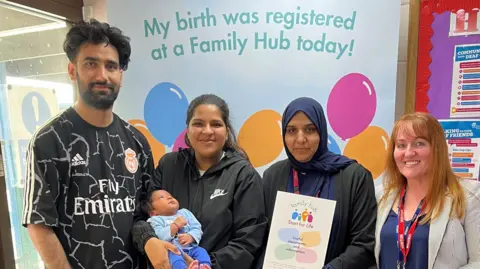 A man and woman pictured holding a baby wearing blue, alongside two other women, one holding a family hub information pack.
