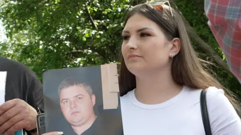 A woman in a white T-shirt and brown hair holds a photo of a young man
