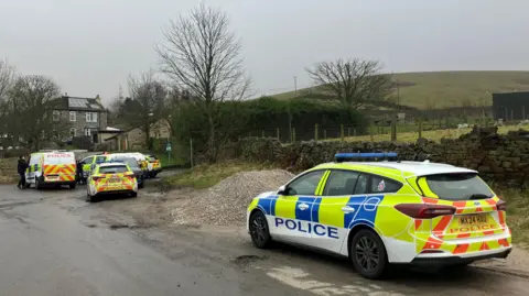 Police cars parked in a country lane close to a dry stone wall. Trees and fields can also be seen.