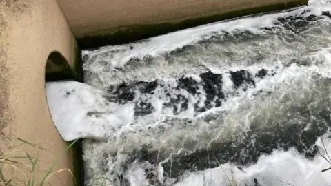 Treated sewage being discharged into a river. The foaming water comes out of a light brown coloured concrete wall with a circular hole in it and lands in waters which also foams.