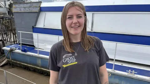 Luke Deal/BBC A woman with long brown hair wearing a grey T-shirt stands on a dock next to a boat. She is smiling at the camera. The boat behind her is white with some blue panels. 