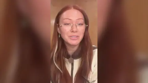 Elz The Witch has long red hair and is wearing rimless glasses and a cream jacket.