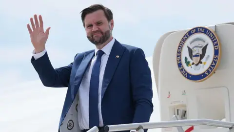 JD Vance waving as he steps out of a jet with the presidential seal on the door. He wears a blue suit with a white shirt and blue tie, and has a pin on his lapel with the US flag on it. 