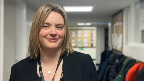 Karen Whitaker. She has shoulder-length blonde hair and is dressed in a black blazer. She is smiling at the camera. She is standing in a school corridor decorated with noticeboards.