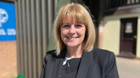 Jackie Gardiner has a shoulder length blonde bob haircut, She is wearing a black jacket and black dress underneath.