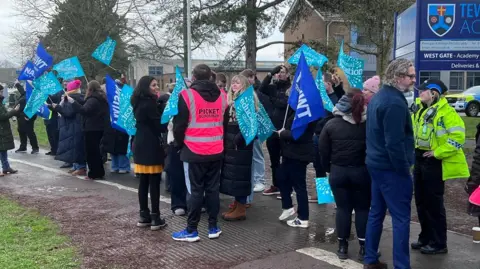 A large group of staff gathers outside Tewkesbury Academy on a dreary February morning. They are wrapped up warm and holding blue union flags as they stand on the pavement and cycle path. A female police officer is stood among them, as are picket supporters wearing pink hi-vis tabards.