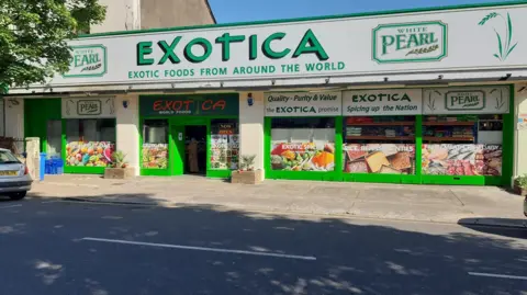 The front of an independent supermarket is shown in green and white colours. Written on it is "Exotica" and "Exotic foods from around the world". In front of it is a pavement, and parked cars.
