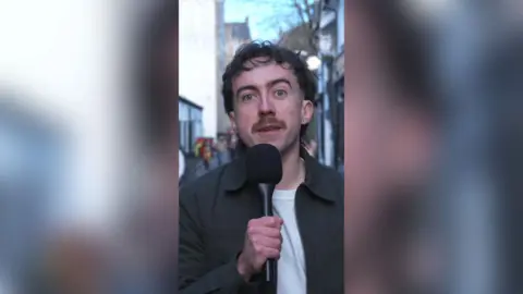 Jack Doherty standing in Glasgow's Ashton lane holding a black microphone. He has short dark hair, a moustache and is wearing a dark grey collared jacket and a white t-shirt.