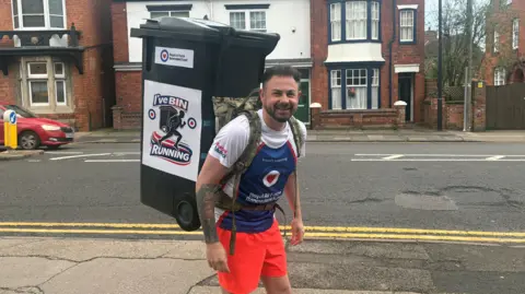 A man dressed in bright orange shorts and a red, white and blue T-shirt. He has a camouflage harness on his back with a large black wheelie bin attached to it. He is standing on a path in front of a road, smiling at the camera. A row of houses is in the background.