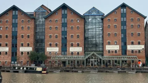 A row of large red brick warehouses with lots of windows, next to a canal. There is a canal boat moored at the front. 