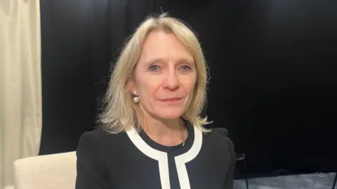 BBC Deputy Elaine Millar is wearing a black and white blazer. She is sat in front of black and beige curtain. 