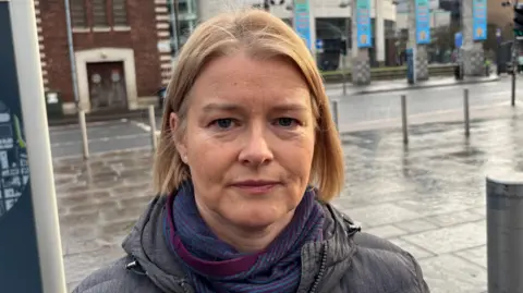 A woman with light blonde hair is standing in a city centre street. Behind her is a wide stretch of pavement and a road. She is wearing a dark grey jacket and a dark blue and purple scarf.