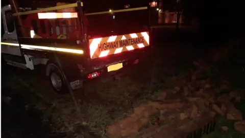 A white van marked "highways maintenance" in front of a pile of bricks. It is night-time. Some of the bricks are on the grass after the wall was hit by the van