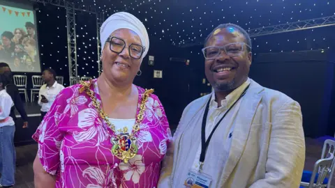 ISCRE Pat Bruce-Browne stands alongside a man. She is wearing a pink flowery dress and gold ceremonial chain around her neck. The man is wearing a beige linen jacket and white shirt. They both have glasses on and are standing in a hall with a stage behind them. 