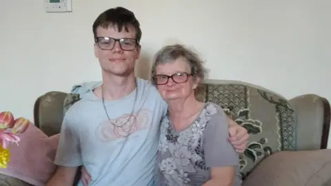Two people sit close together on a sofa in a living room, with drinks and snacks on a table in front of them.
