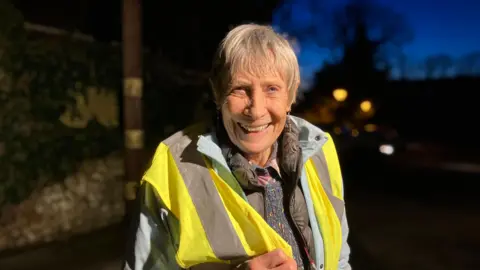 Emily Coady-Stemp/BBC Penny Aeberhard looks at the camera and is wearing a high vis jacket. She is looking at the camera and smiling and has short grey hair.