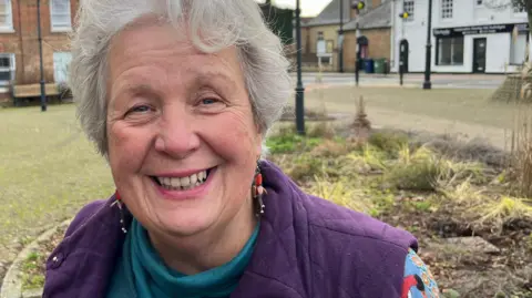 John Devine/BBC Sue Unwin has short grey hair and is smiling. She is wearing a turquoise jumper and a purple gilet. Behind her is a small garden, black lampposts and buildings. 
