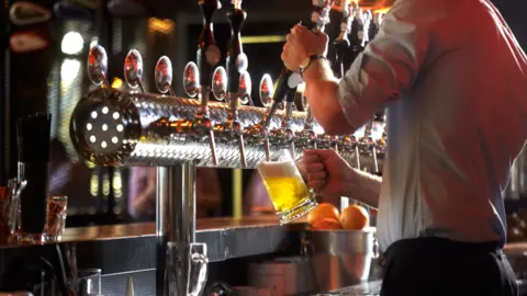 Getty Images A gray-shirted bar worker in a pub pulls a pint of lager from the beer taps with a silver bucket of oranges in front of him