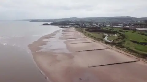 A drone shot from above Dawlish Warren beach in Devon. You can see the sea, beach, a large green area and homes and buildings in the distance. 