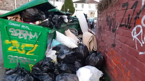 A row of large, green municipal bins surrounded by black big bans full of rubbish blocking the pavement.