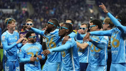 Sky Blues players, including Victor Torp, Jay Dasilva, Frank Onyeke, Brandon Thomas-Asante, Joel Latibeaudiare and Haji Wright celebrate winning the EFL Championship title. They are wearing the club's home kit, with some wearing shirts celebrating the occasion.