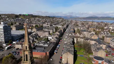 Aerial view of housing in Inverclyde, with a long straight street lined by tenement buildings, a church spire in the foreground and the River Clyde visible in the distance. 