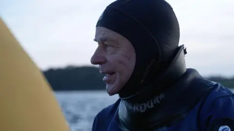 BBC A man wearing a black and blue diving suit. He is looking to the right. There is water dripping from his chin.