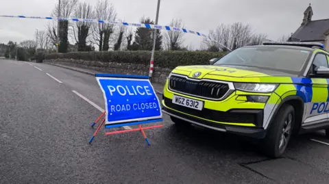 BBC A police SUV next to a sign saying "police, road closed" and cordon police tape blocking the road. there is a stone wall, with a hedge running down the road. 