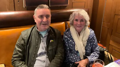 A man wearing a top and black jacket sat next to a woman, wearing a blue coat and white scarf. They are both looking at the camera and smiling. There are sat on a light brown seat. The wall behind them is wood. 
