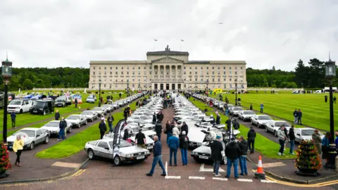 Pacemaker Stormont. In front of the building are multiple rows of parked Delorean cars. There are dozens of people walking around and looking at the vehicles. It is a cloudy day. 