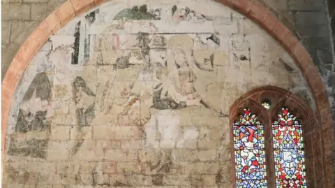 The photo shows the inside wall of a church. On the right is a small stain glass window, while to the left of it can be seen the faded detail of a painting on the wall. The outline of several figures can just about be made out.