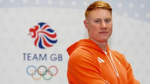Tom Dean, looking at the camera. He has an orange hoodie on, has ginger hair, and is standing in front of the Team GB logo and the Olympic rings