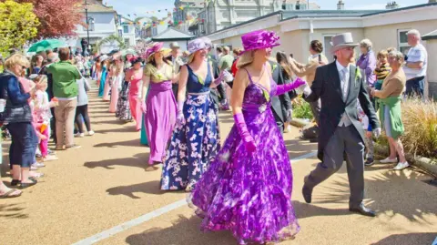Couples are walking along a gravel walkway away from a street with bunting. The ladies are wearing brightly-coloured full-length gowns with hats and long gloves. The gentlemen are wearing top hats and tails. It is bright sunny day and the path is lined with spectators.