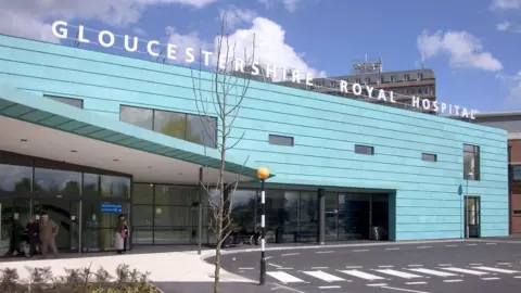 The front exterior of Gloucestershire Royal Hospital. It is a large rectangular building clad in blue panels. On the left there is a sheltered area, and a zebra crossing heading. It is a sunny day with wispy clouds in the sky.