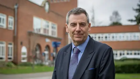 A man with short grey hair is pictured wearing a dark blue suit with a light blue shirt and dotted tie. He is stood in front of a large red brick building with hedges and grass behind him.