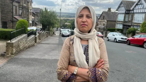 Aisha Iqbal/BBC A woman stands with arms folded in the middle of a residential street. It's a tidy, well-kept area with houses with classical facades and rolling countryside visible in the background. She wears a patterned top and a beige coloured headscarf.