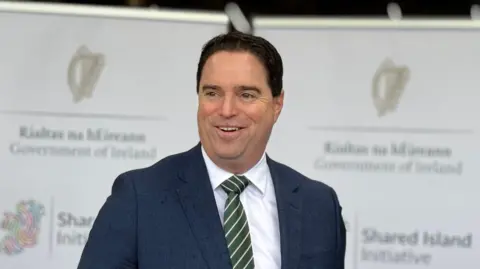 Martin Heydon, un hombre de pelo corto y negro, sonríe durante una rueda de prensa. Lleva una chaqueta azul marino a cuadros, camisa blanca y corbata a rayas verdes y blancas. Detrás de él, una pancarta luce el logotipo del gobierno irlandés y la inscripción «Iniciativa de la Isla Compartida».
