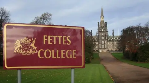 A red sign with "Fettes College" written in gold next to a coat of arms. The central turret of the stone school is visible at the end of a long pathway.