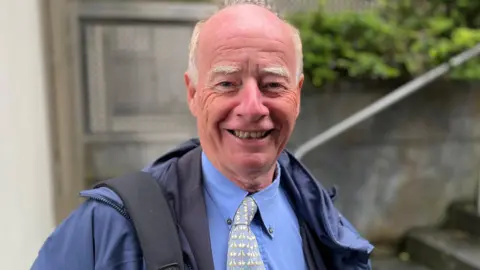 Deputy George Oswald looking towards the camera. He's smiling and has a bald head with white hair on the side of his face.
he is wearing a blue overcoat, with a blue shirt and a blue and white tie.