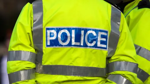 A stock image showing the back of a police officer, in a fluorescent yellow jacket with the word police in white capital letters on a blue background.