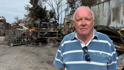 Martin Fey looks at the camera with a neutral facial expression. He's dressed in a blue and white striped polo shirt. There are several burnt vehicles behind him.