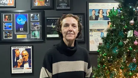 A woman wearing a striped black and grey turtle neck jumper standing next to a Christmas tree on her right and behind her is a black painted wall with different movie posters on it.