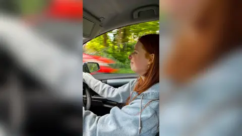 A young female driver with medium length red hair driving in a car. She is wearing a light grey hoody and there are trees visible out the driver's window and a passing red car.