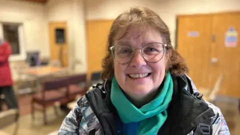 Shariqua Ahmed/BBC Sandra, a woman with blonde hair, wearing a green scarf and a multi-coloured jacket, smiling for camera. 